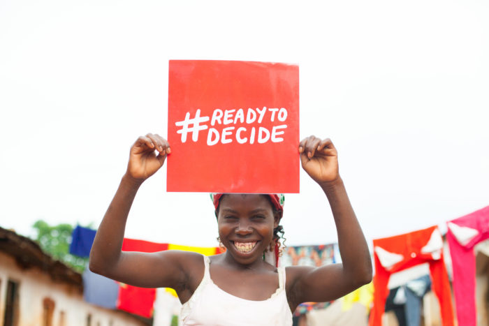 A young woman smiles and holds up a poster saying Ready To Decide