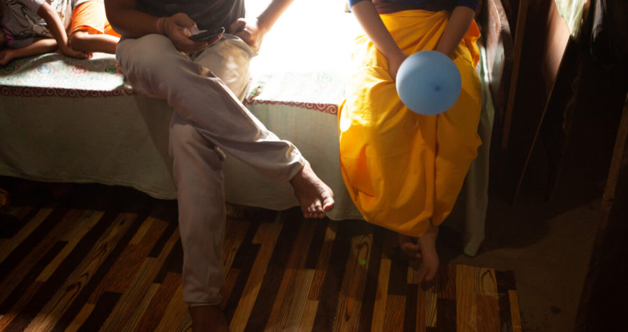 Two people sit on a bed, one holds a mobile phone and the other holds a balloon