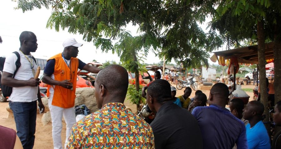 A group of people listen to a man wearing an orange vest