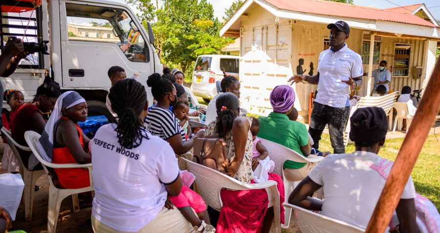 A man standing outside and wearing a baseball cap speaks to women sitting in chairs in Uganda