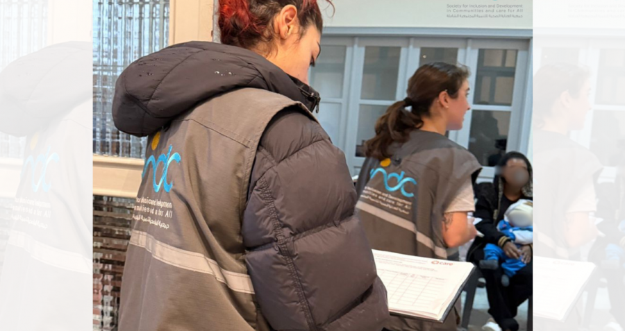 Two women stand with clipboards. A man sits on a bench in front of a building with a young child on his lap.