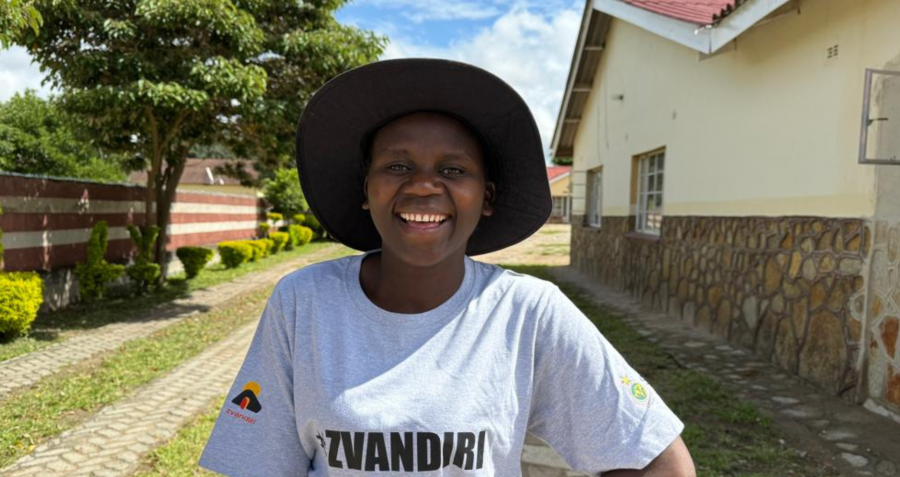 A young black woman wearing a t-shirt and a hat stands in front of a house.
