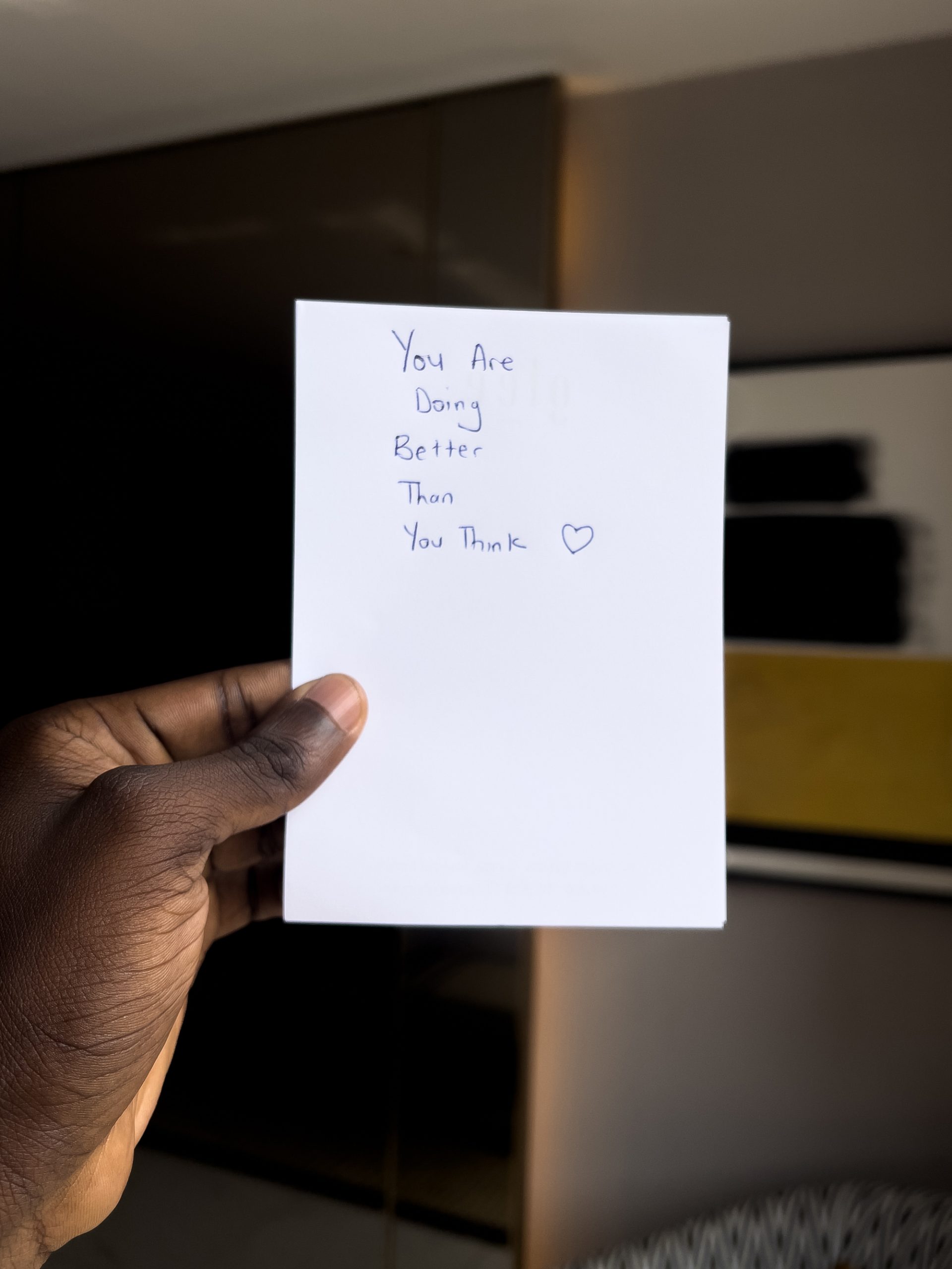 A handwritten note on a blank piece of paper held by a person with a black hand reads: "You are doing better than you think" with a small hand drawn love heart to the right