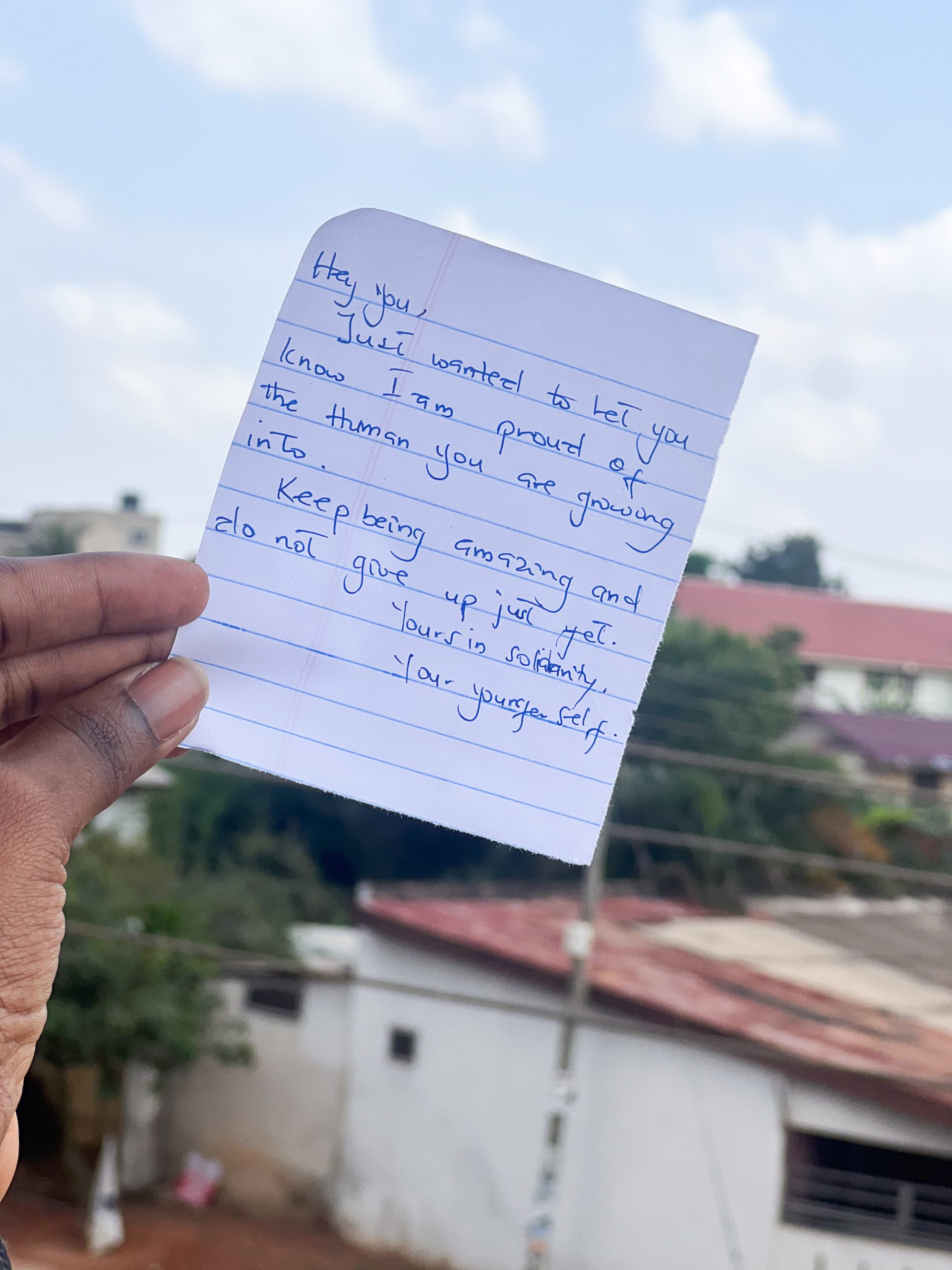 A note, held by a black in the air against a backdrop of buildings and trees reads: “Hey you, just wanted to let you know I am proud of the human you are growing into. Keep being amazing and do not give up just yet.”